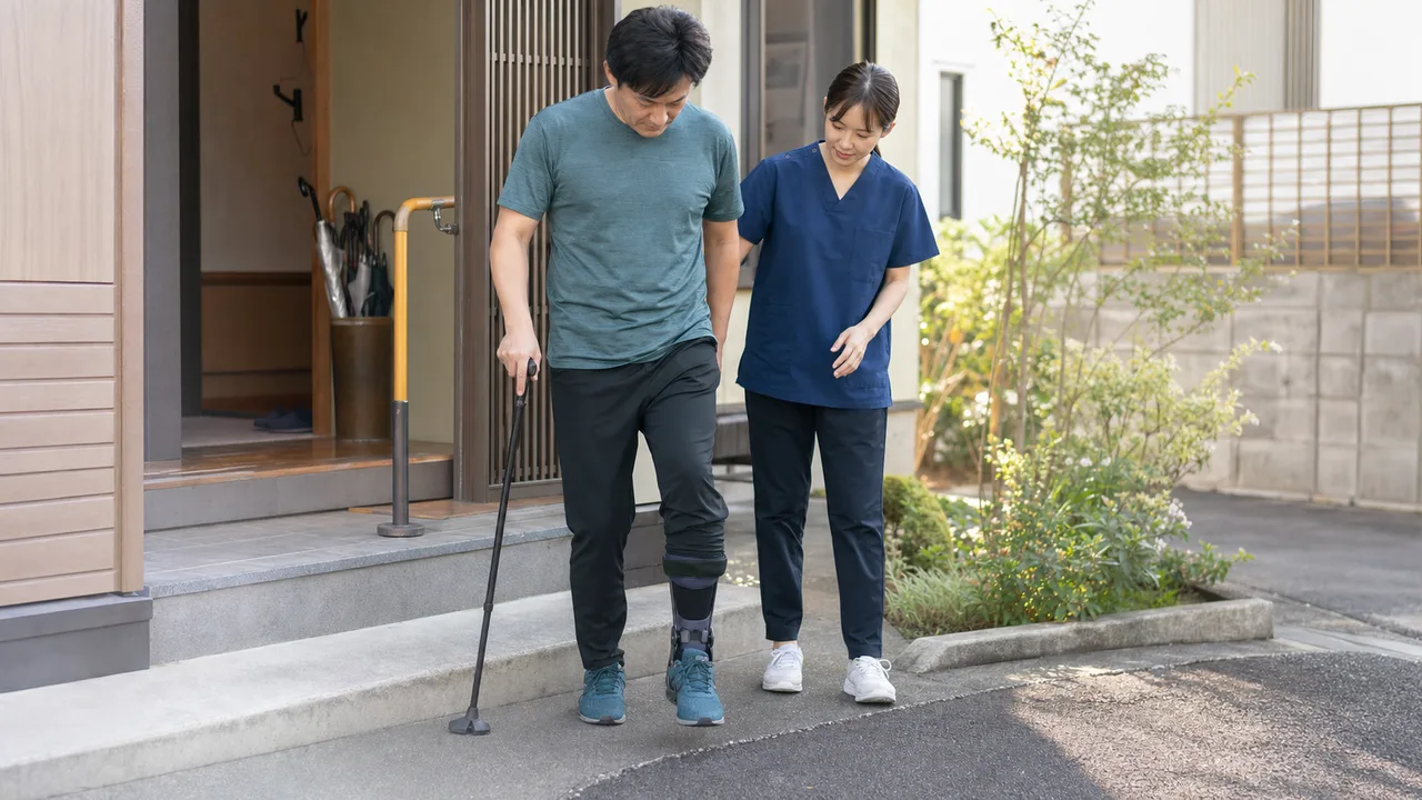 自宅玄関の段差で杖を使い歩行練習を行う人と付き添うリハビリ専門職